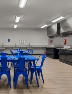 Cafeteria with bright blue metal tables and chairs, and a stainless steel kitchen area along the back wall.