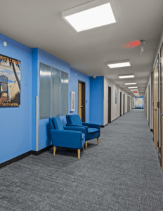 Indoor hallway with blue walls and a small seating area of two blue chairs against the left wall, gray carpet, and bright ceiling lights at intervals.