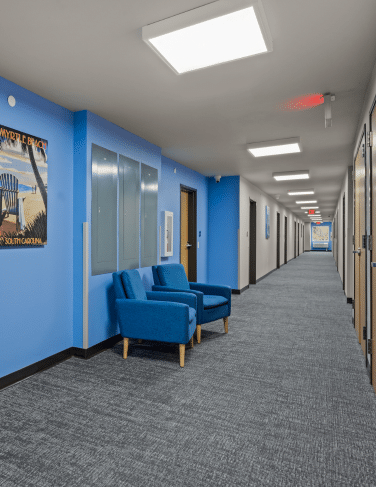Indoor hallway with blue walls and a small seating area of two blue chairs against the left wall, gray carpet, and bright ceiling lights at intervals.