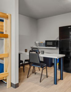 Dorm room kitchenette with a white table and two chairs, microwave, coffee maker, toaster, full-height fridge, and a wooden bunk bed beside.